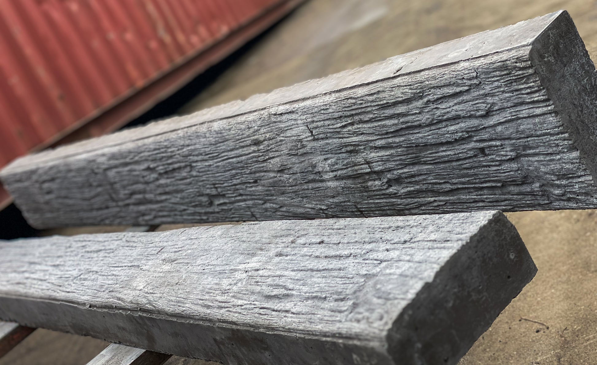 Two weathered gray wooden concrete sleepers with a realistic bark-like texture, stacked on a surface with a red corrugated metal container in the background.